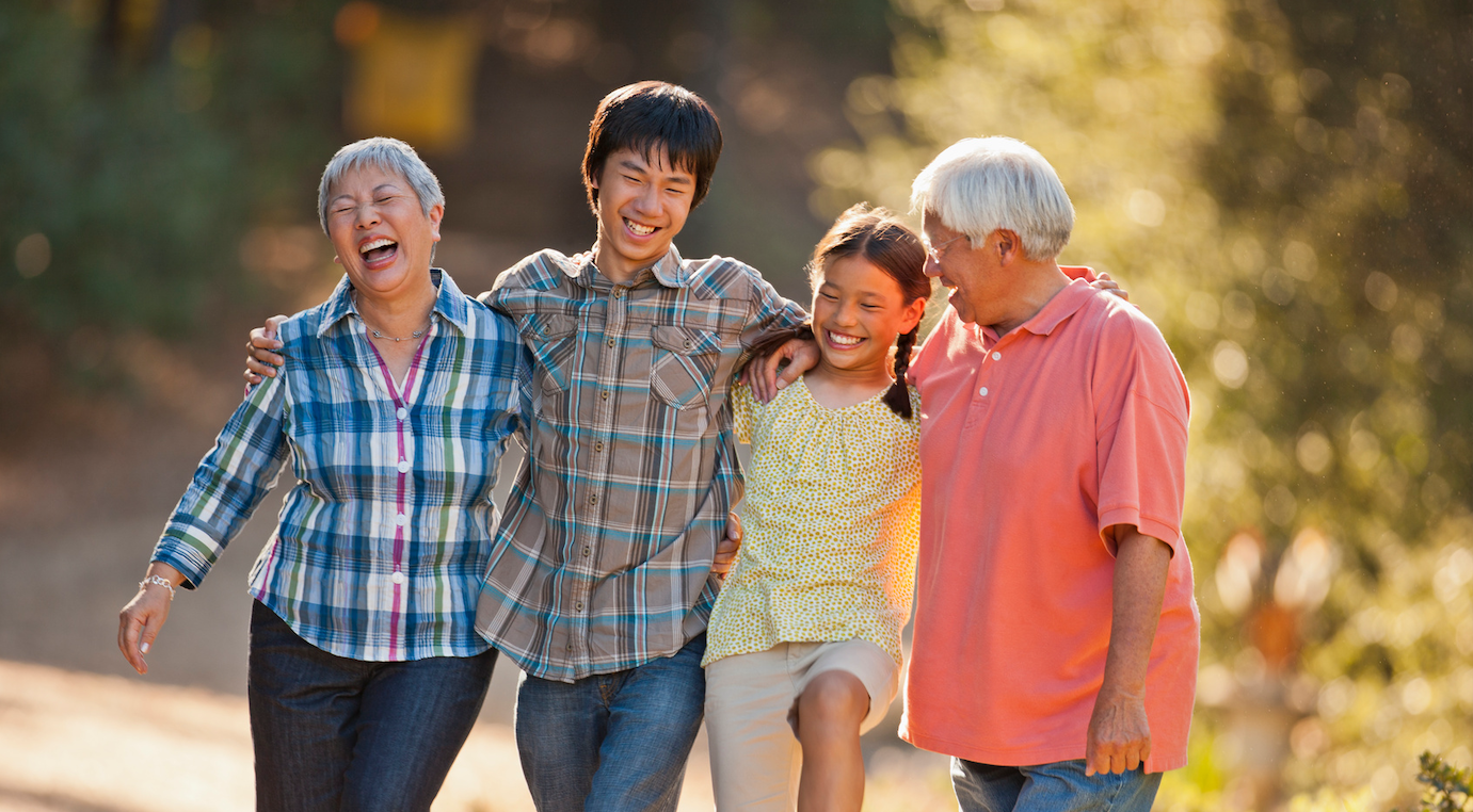 Four people, two older adults and two children, walk outdoors arm in arm, laughing and smiling together in a sunlit, natural setting.